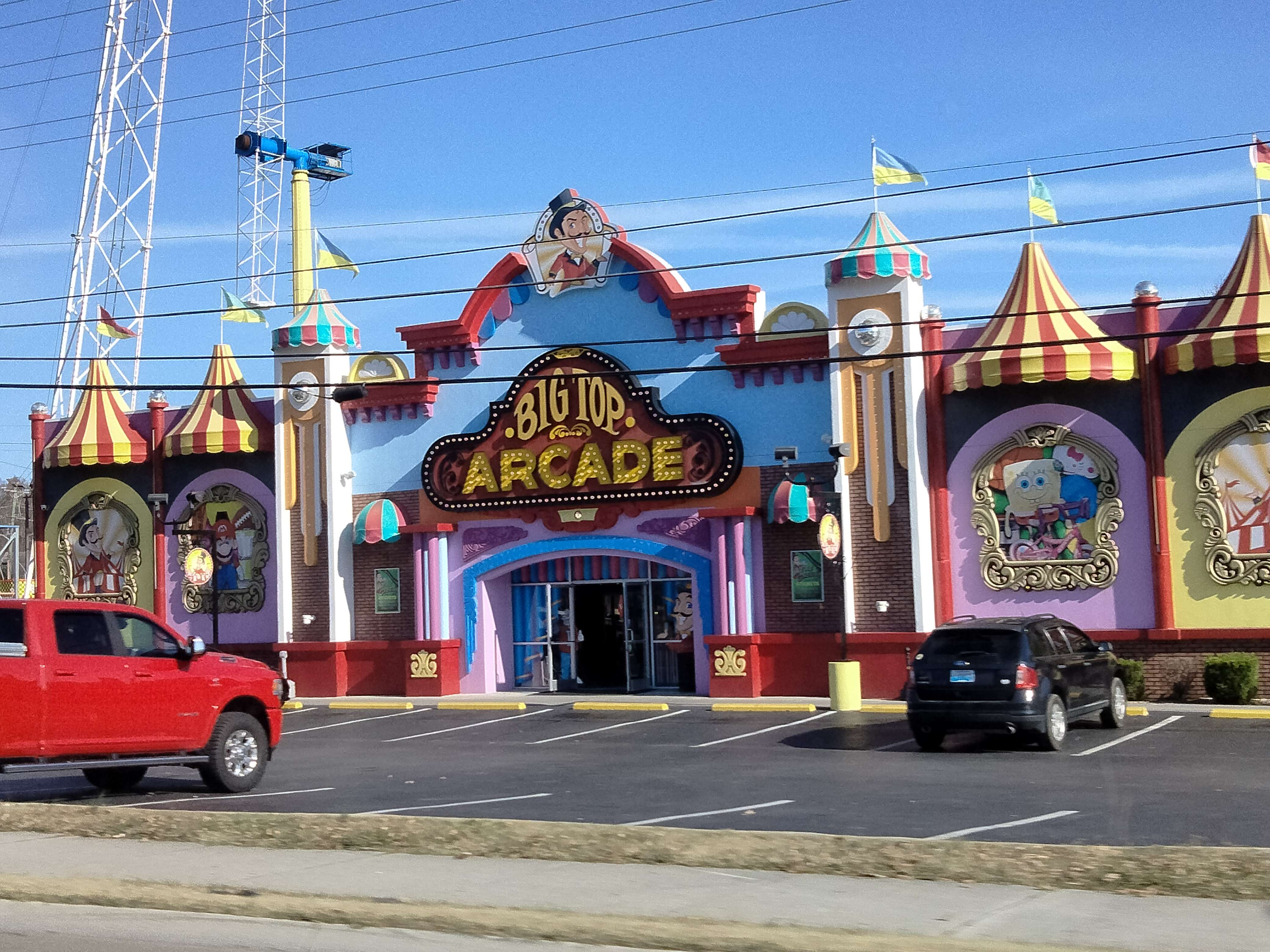 A picture of a roadside arcade taken from the perspective of someone sitting in the passenger seat of a car. The arcade is circus-themed and called Big Top Arcade with extravagantly themed architecture in primary colors.