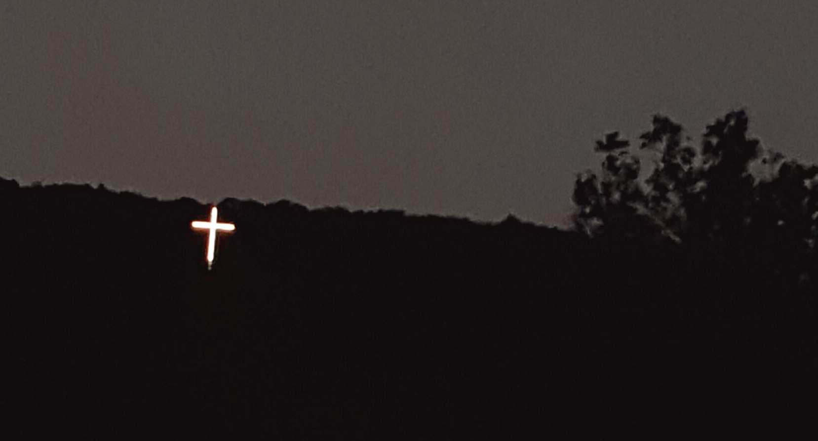 A picture of a silhouetted mountainscape at twilight. A bright cross is illuminated against the dark.