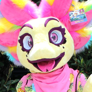 A photograph of an axolotl fursuiter from the head up. She is looking to the camera and has a happy expression. She has pastel yellow skin with pink gills and rainbow accents.